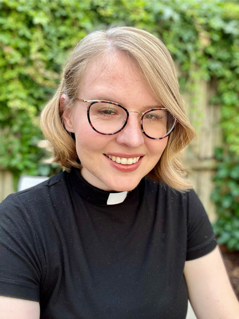 Pastor Madeline Tallman, wearing clerical, looking toward camera, with vegetation background.